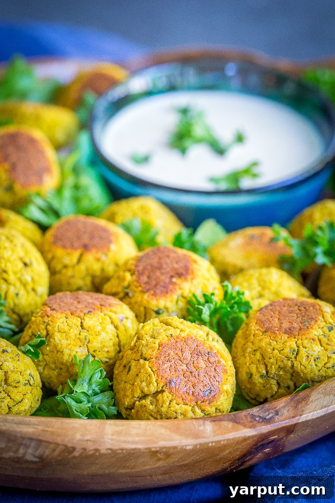 A bowl of creamy, light-colored tahini dipping sauce alongside a beautifully arranged plate of golden-brown roasted butternut squash falafel bites, garnished with fresh herbs.