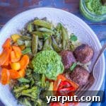 A vibrant bowl filled with roasted broccoli, carrots, snap peas, and bell pepper, alongside quinoa and Gardein meatless meatballs, all drizzled with a fresh spinach mint pesto, perfect for a healthy spring meal.