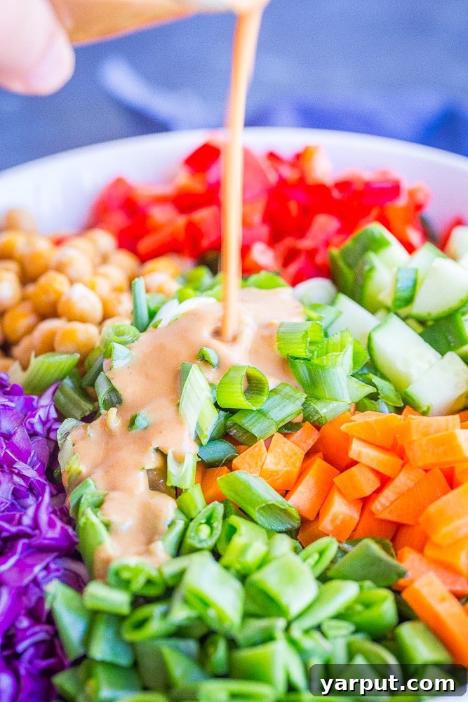 The creamy ginger cashew dressing being generously poured from a glass jar over a fresh, colorful Asian Chickpea Chopped Salad in a large mixing bowl, ready to be tossed.