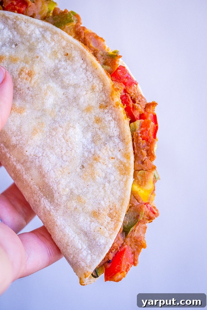 Baked Tacos Bursting with Summer Vegetables 4 A hand holding a crispy baked taco, showcasing the delicious filling of refried beans and sautéed summer vegetables spilling out slightly, highlighting its texture.