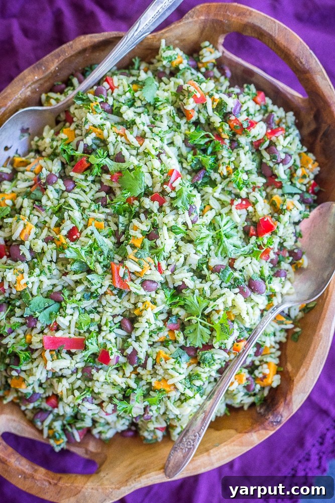 A large bowl of bright green rice mixed with black beans and orange sweet potato cubes, garnished with fresh herbs, with two serving spoons ready for dishing.