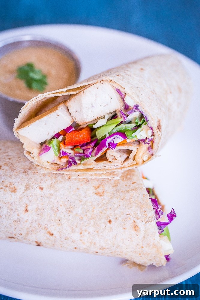 A Crunchy Asian Tofu Peanut Wrap, expertly sliced in half, rests on a plate alongside a small bowl of creamy peanut sauce, ready for dipping.