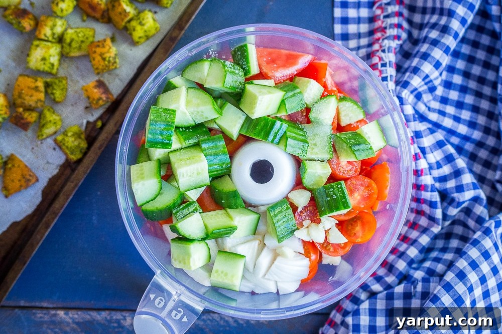 A food processor bowl filled with colorful chunky cherry tomato gazpacho ingredients, alongside a baking sheet loaded with homemade mint pesto croutons ready for the oven.