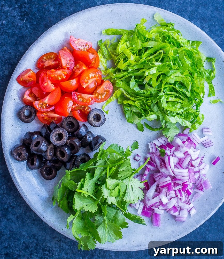 Fiesta Pizza Bagels 4 Various Taco Pizza Bagel toppings like black olives, lettuce, tomato, and avocado arranged on a white plate