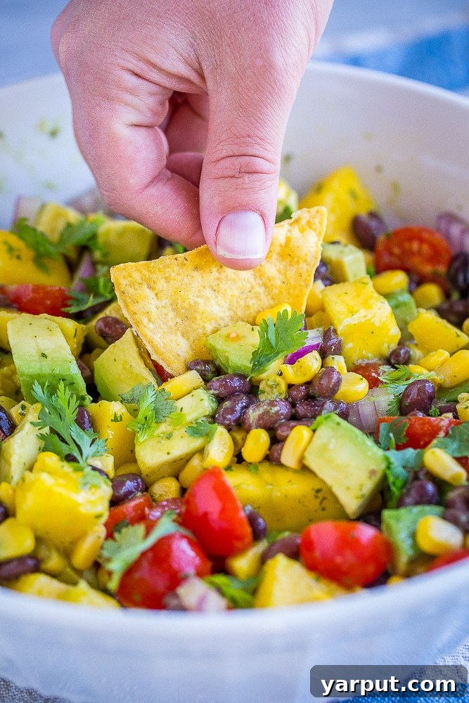 A hand dipping a crispy tortilla chip into the colorful black bean salad with mango and avocado, showcasing its appeal as a snack or appetizer.