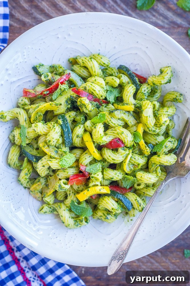 Vibrant Summer Pesto Pasta 2 A large, inviting bowl of Pesto Pasta featuring bright green pesto coating al dente pasta, generously mixed with colorful summer vegetables like golden-brown zucchini, yellow summer squash, and red bell peppers, garnished with fresh basil leaves.