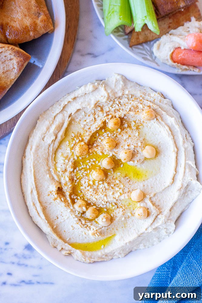 The Secret to Perfectly Creamy Hummus 2 A beautifully presented bowl of creamy hummus, garnished with a drizzle of olive oil, a sprinkle of paprika, and fresh parsley. It is surrounded by warm pita bread and a vibrant assortment of fresh vegetables like cucumber sticks, carrot sticks, and bell pepper slices, inviting a delightful dip.