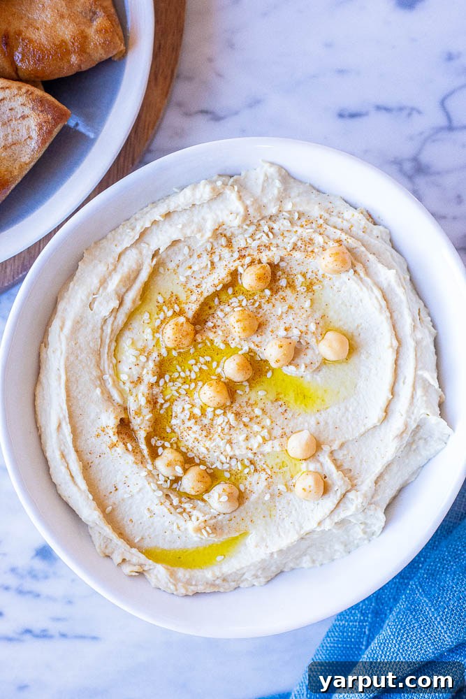 The Secret to Perfectly Creamy Hummus 5 The creamiest hummus recipe presented in a rustic bowl, accompanied by golden pita bread triangles and fresh green garnishes. The texture is impeccably smooth, promising a luxurious dining experience.