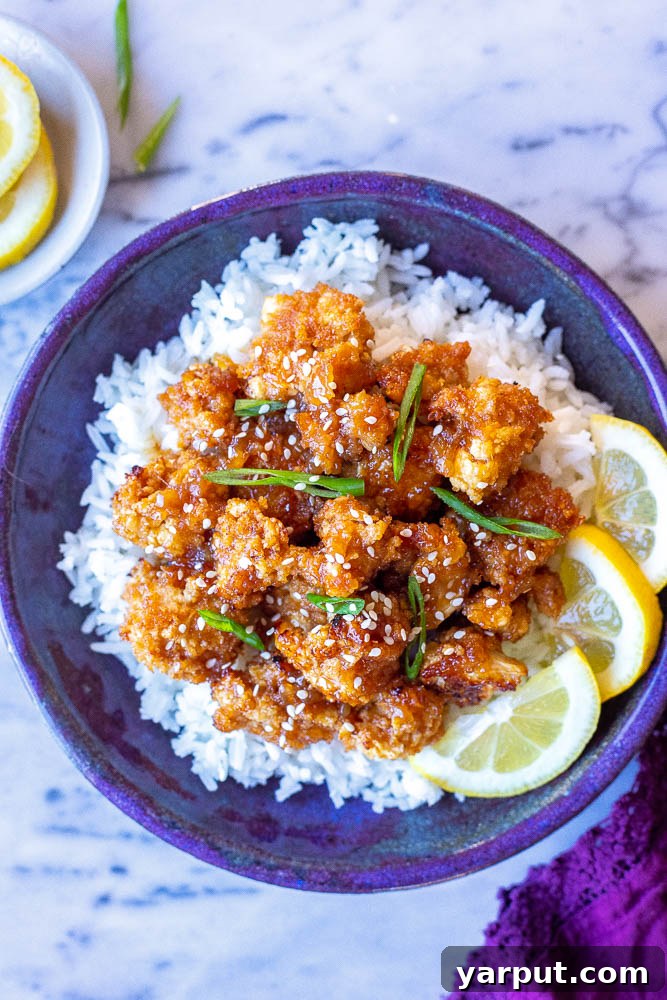 Lemon-Glazed Roasted Cauliflower 2 A beautifully presented bowl of crispy sticky lemon cauliflower served over a bed of fluffy white rice, garnished with fresh green onions and sesame seeds. The vibrant yellow sauce glistens on the cauliflower florets.