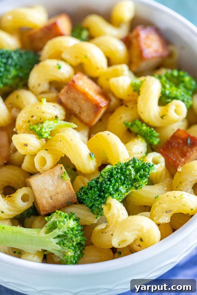 Close-up photograph of a single serving bowl of Tofu Broccoli Pasta, beautifully presented and garnished with a sprig of fresh herbs