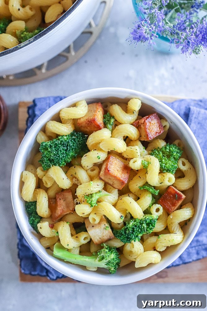 A beautifully arranged bowl of Tofu Broccoli Pasta, with a fork nestled in, placed on a rustic wooden table alongside a textured napkin, highlighting its appetizing appearance.