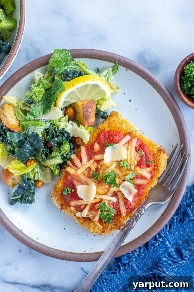 Cheesy Vegan Tofu Parmesan 7 Vegetarian chicken parmesan on a white plate, served alongside a fresh green salad, with a fork ready for dining.