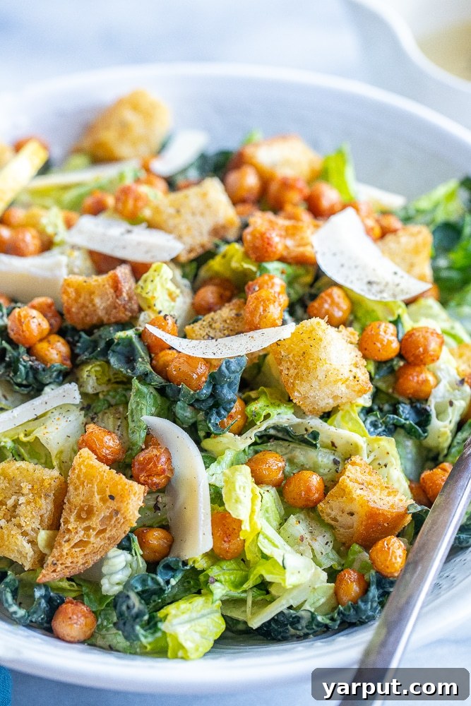A close-up view of a perfectly tossed vegan Caesar salad in a rustic bowl, showcasing the creamy dressing coating each leaf of romaine and kale, with visible croutons and chickpeas.