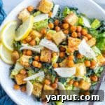 Close-up of a serving of vegan Caesar salad, showing the croutons, chickpeas, and vegan parmesan cheese. It is served with fresh lemon slices.