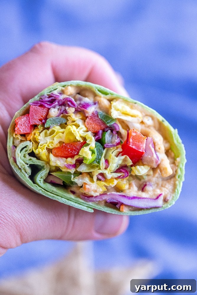 Plant-Powered Peanut Hummus Wraps 6 A hand holding a vibrant rainbow veggie hummus wrap, showcasing the fresh and colorful ingredients packed inside, against a blurred background.