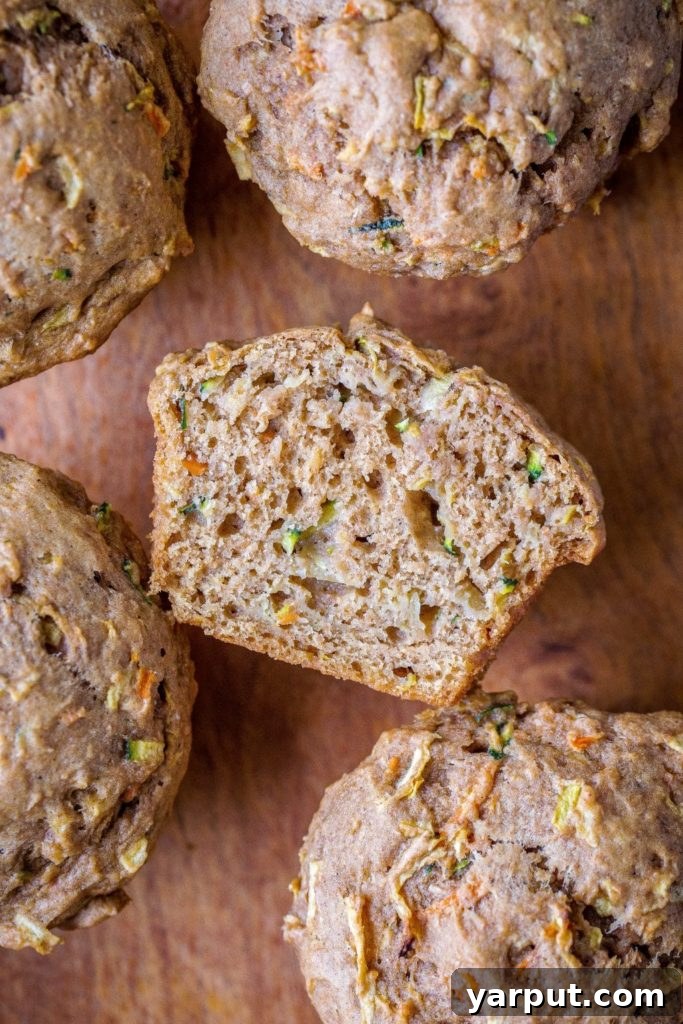 Farmhouse Zucchini Apple Carrot Muffins 2 Healthy Zucchini Apple Carrot Muffins on a wooden cutting board, with one muffin cut in half to reveal the vibrant ingredients inside.