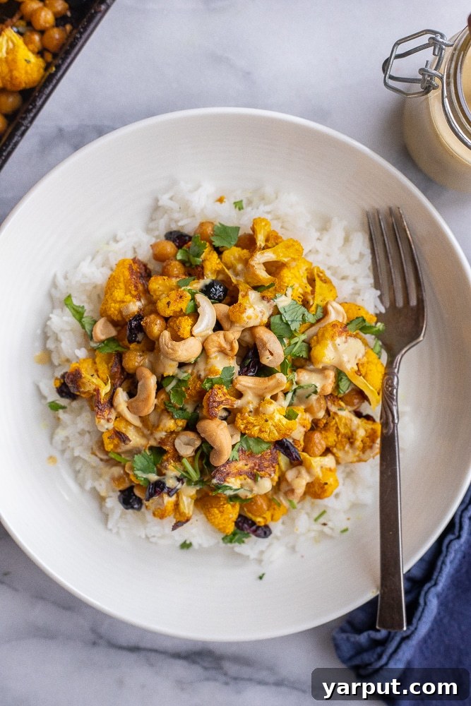 An overhead shot of a bowl filled with golden roasted cauliflower, chickpeas, and dried cranberries, generously drizzled with tahini dressing.