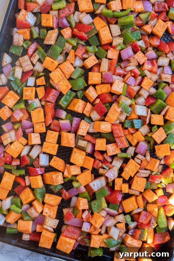 Golden Sweet Potato Skillet 3 Sweet potato hash ingredients laid out on a sheet pan before roasting, including diced sweet potatoes, red and green bell peppers, and red onion, all coated lightly in olive oil and spices, ready for the oven.