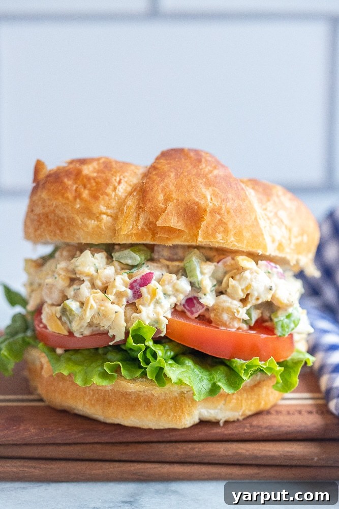 Close up of a golden, flaky vegetarian tuna salad croissant filled with a creamy chickpea mixture, lettuce, and tomato, resting on a rustic cutting board. This plant-based sandwich is ideal for a healthy and satisfying lunch, perfect for picnics or summer adventures.