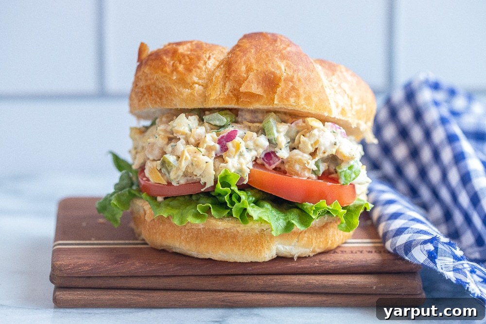 A perfectly assembled vegetarian tuna salad croissant sandwich, featuring a rich chickpea salad, fresh green lettuce, and vibrant red tomato slices, presented on a plate. This image showcases the appealing final product, ready for a healthy meal.
