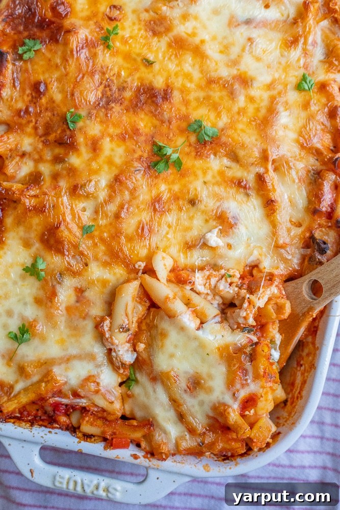 A large pan of freshly baked vegetarian ziti, golden brown and bubbling with melted mozzarella cheese, ready to be served with a wooden spatula.
