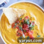 A small bowl of creamy vegan pumpkin queso dip with a chip on the side.