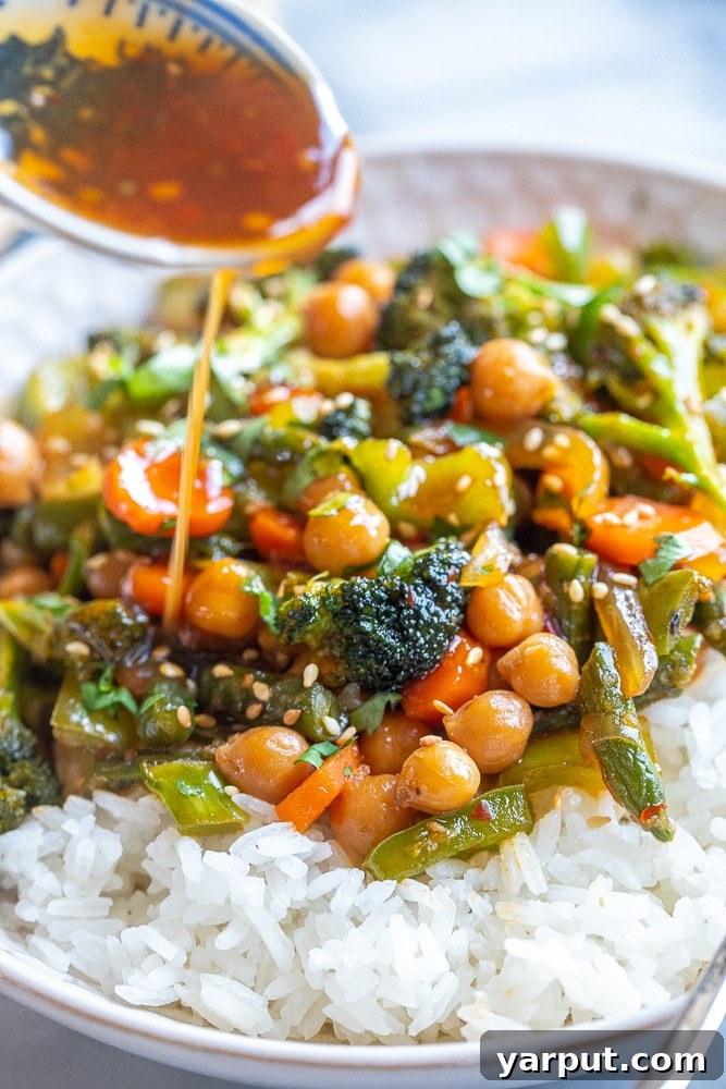 A spoon pouring glossy chili garlic sauce over a fresh bowl of stir fry vegetables with chickpeas, highlighting the rich texture of the sauce.