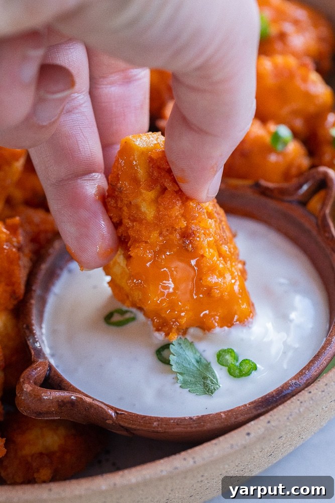 Irresistible Vegan Buffalo Tofu Wings 6 A hand dipping a perfectly golden and saucy vegan buffalo tofu wing into a creamy white vegan ranch dressing, highlighting the delicious texture and vibrant color.