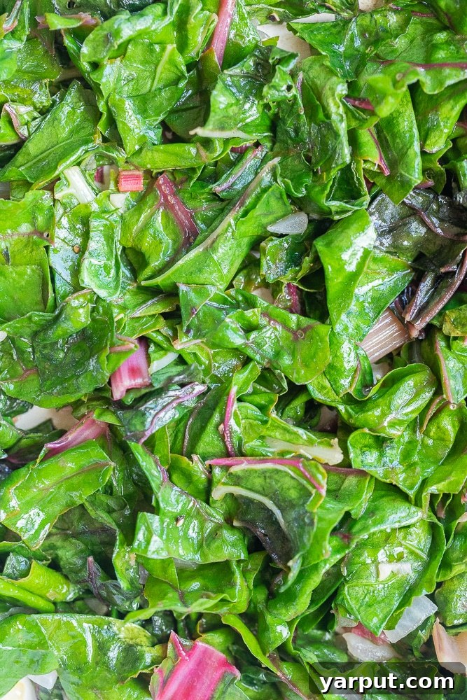 Effortless Swiss Chard Sauté 6 A close-up view of fresh Swiss chard leaves and stems cooking in a hot pan with aromatics, showcasing the wilting process and vibrant colors.