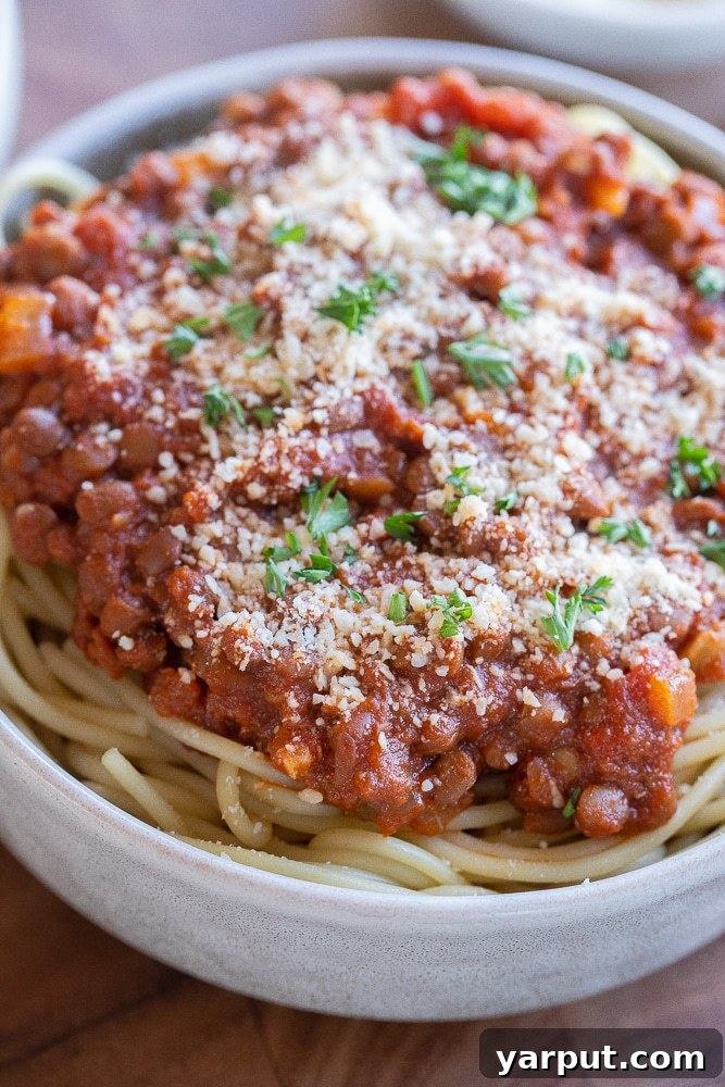A close-up of a single bowl of meaty lentil spaghetti, generously topped with freshly grated Parmesan cheese and vibrant green parsley, highlighting the rich texture of the sauce.