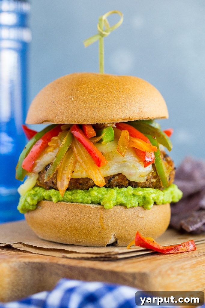 Fajita Baharatlı Kara Fasulye Burger 4 Close-up of a fully assembled Black Bean Fajita Veggie Burger, featuring a crispy patty, melted pepper jack cheese, vibrant peppers and onions, and guacamole on a toasted bun.