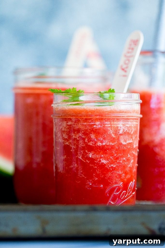 Sadece Karpuzla Buz Gibi Serinlik! 4 A person's hand holding a glass of vibrant watermelon slushie, with more watermelon in the background.
