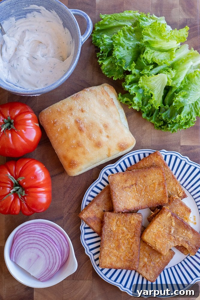 All the fresh ingredients laid out for making vegan BLT sandwiches: bread, lettuce, tomatoes, tofu bacon, and a bowl of lemon dill mayo.